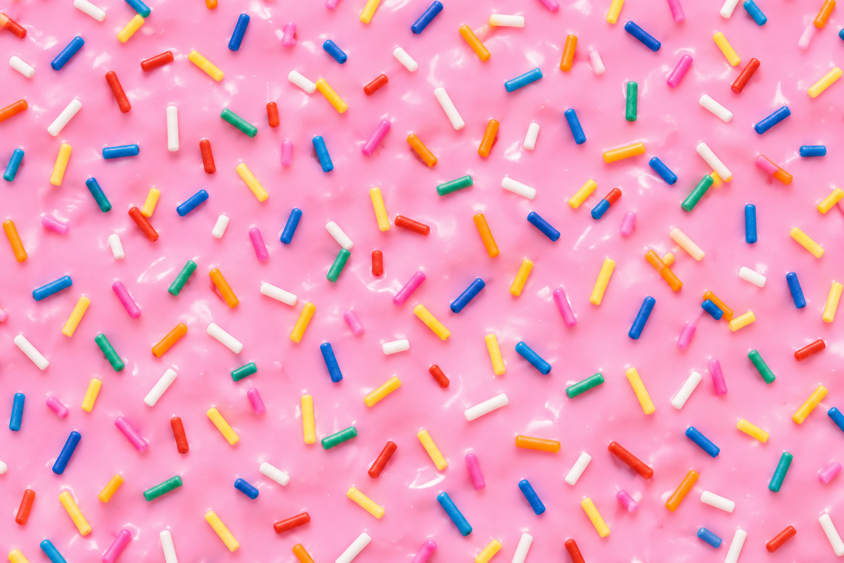 A close-up of pink frosted candy canes or sprinkles, covered with colorful cylindrical sprinkles in blue, yellow, orange, green, red, and white.