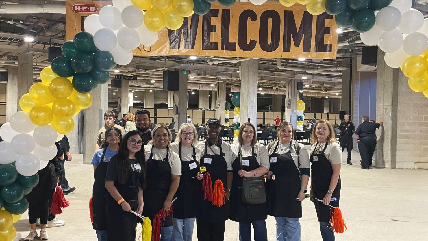 Group of people in aprons posing under a 'WELCOME' sign with balloons in a large indoor space.